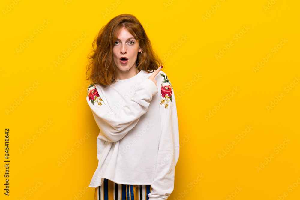 Young redhead woman over yellow wall surprised and pointing side Young redhead woman over yellow wall surprised and pointing side