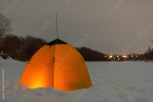 Fishing yellow winter tent is glowing with the light of candles inside with the shadow of a fisherman on the snowy ice of the river near the forestof the city lights in the distance.