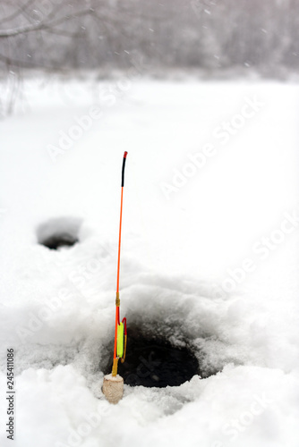 Homemade winter fishing rod stands at the hole in the ice of the river against the snow-covered forest. photo