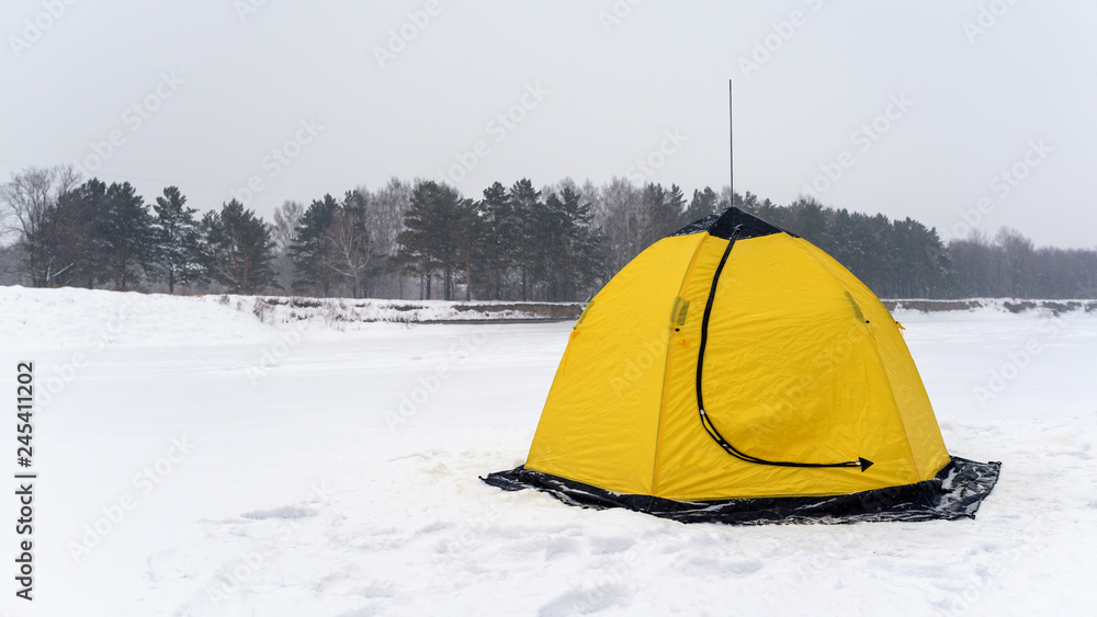 Yellow fishing winter tent in the form of a Lotus stands on the snowy ice of the river closed against the snow-covered forest trees in winter.