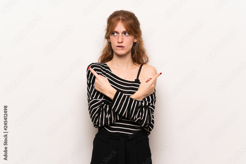 Young redhead woman over white wall pointing to the laterals having doubts Young redhead woman over white wall pointing to the laterals having doubts