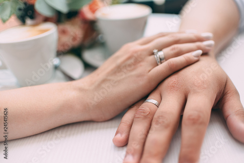 The bride and groom hold hands, holding expensive wedding rings with white gold, on the background of a wedding bouquet and two cups of coffee on a white table.
