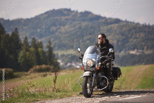 Handsome bearded motorcyclist with long hair in black leather jacket and sunglasses sitting on cruiser motorcycle on roadside on blurred background of empty twisty road on sunny day.