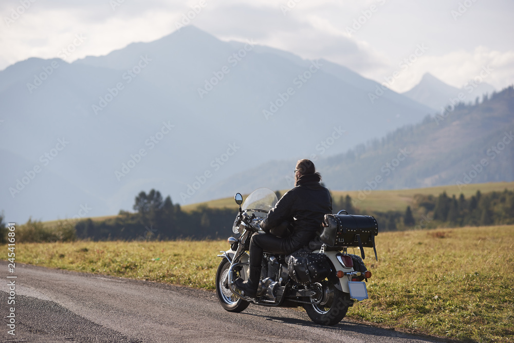 Back view of biker in black leather jacket riding cruiser motorcycle along road on blurred copy space background of beautiful foggy mountain range and cloudy sky on bright sunny day.