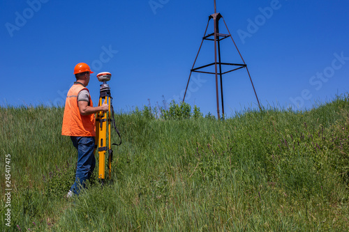 Professional Male Land Surveyor Measures Ground Control Point Using a GPS Rover. Green Field on a Background.