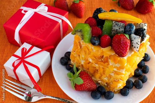 Beautiful  delicious  fruit cake in white plate  red and white gift boxes  fork on orange tablecloth.