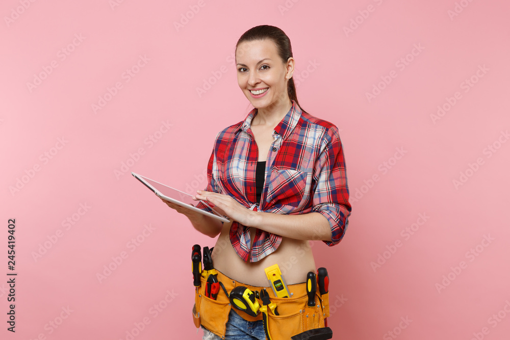 Young handyman woman in plaid shirt, denim shorts, kit tools belt full of variety instruments hold tablet pc computer money isolated on pink background. Female doing male work. Renovation concept Young handyman woman in plaid shirt, denim shorts, kit tools belt full of variety instruments hold tablet pc computer money isolated on pink background. Female doing male work. Renovation concept