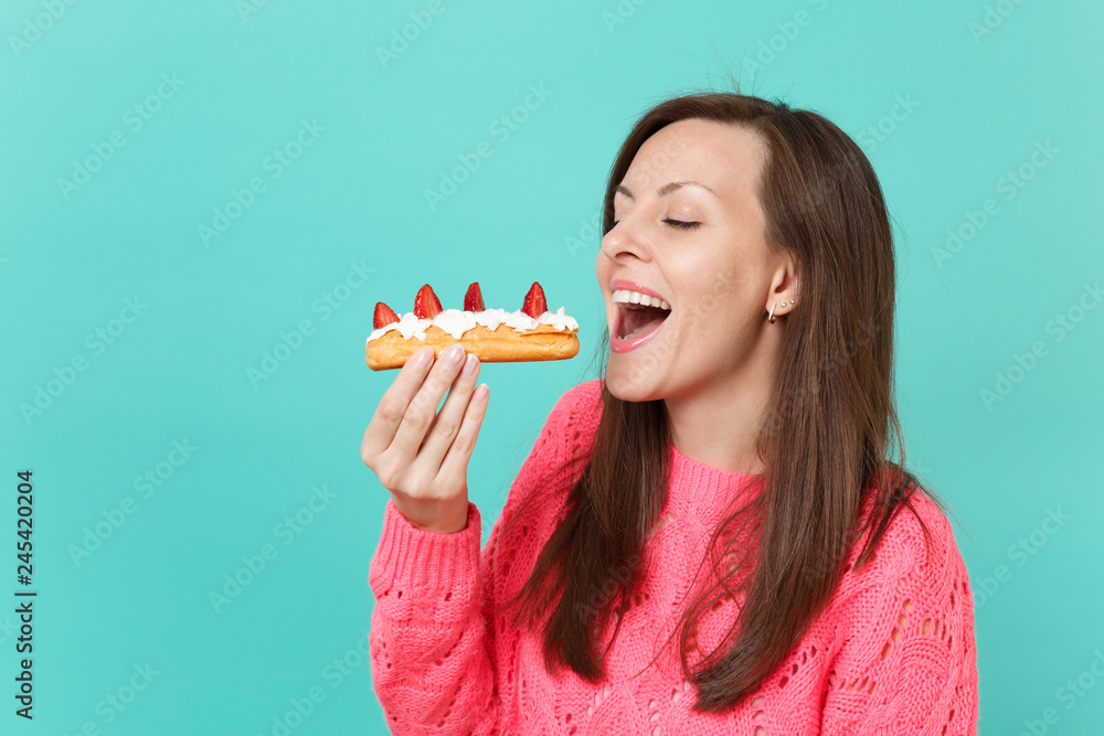 Φωτογραφία Stunning joyful young woman in knitted pink sweater holding in hand and eating eclair cake isolated on blue turquoise wall background, studio portrait