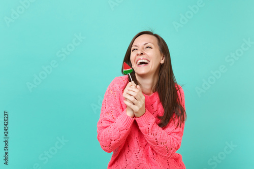 Laughing funny young woman in knitted pink sweater looking up, holding watermelon lollipop isolated on blue turquoise wall background, studio portrait. People lifestyle concept. Mock up copy space.