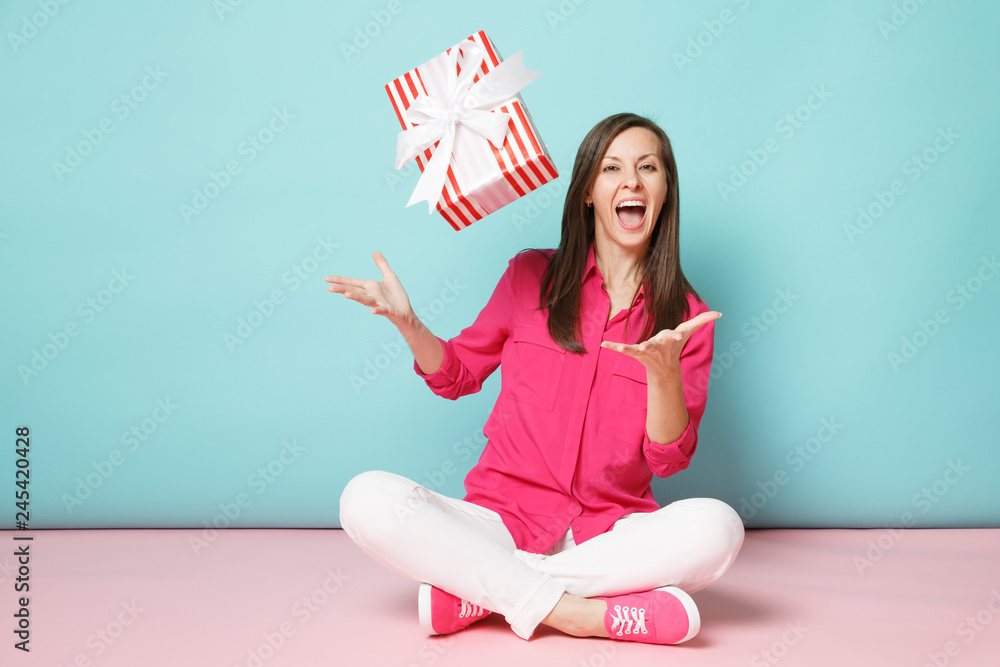 Full length portrait woman in rose shirt blouse white pants sitting on floor hold present box isolated on bright pink blue pastel wall background studio. Fashion lifestyle concept. Mock up copy space. Full length portrait woman in rose shirt blouse white pants sitting on floor hold present box isolated on bright pink blue pastel wall background studio. Fashion lifestyle concept. Mock up copy space.