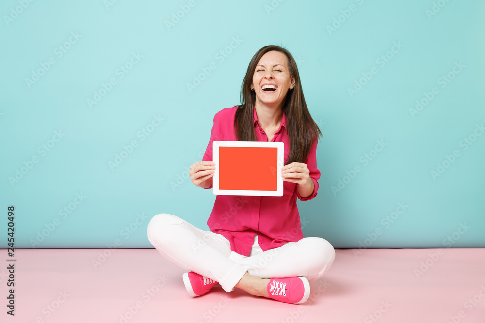 Full length portrait woman in rose shirt blouse, white pants sitting on floor hold tablet pc isolated on bright pink blue pastel wall background studio. Fashion lifestyle concept. Mock up copy space. Full length portrait woman in rose shirt blouse, white pants sitting on floor hold tablet pc isolated on bright pink blue pastel wall background studio. Fashion lifestyle concept. Mock up copy space.