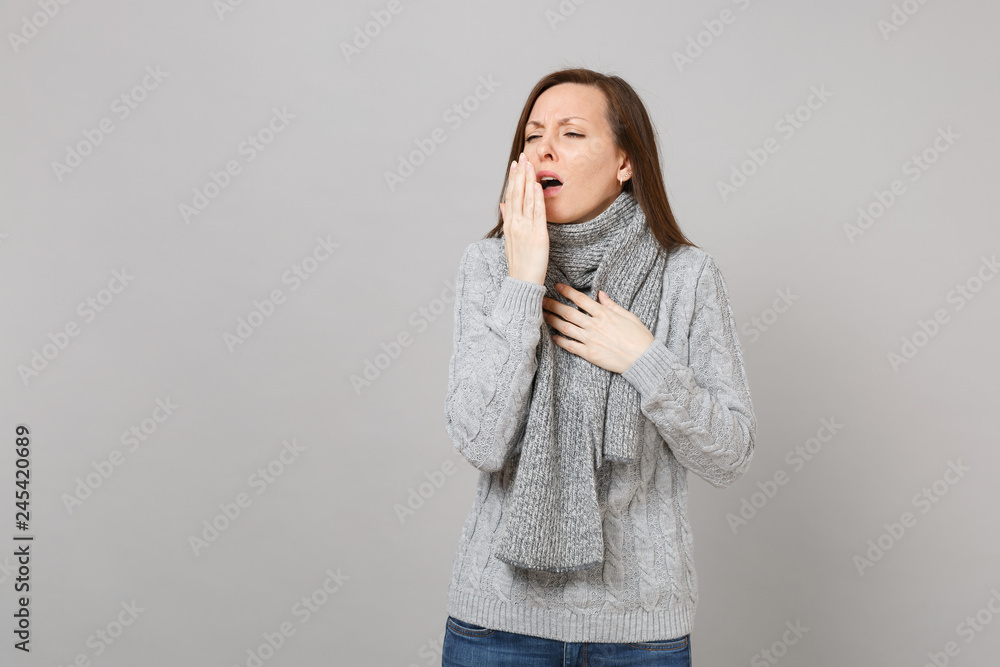 Young woman in gray sweater scarf sneezing or coughing covering mouth with palm isolated on grey wall background. Healthy lifestyle ill sick disease treatment, cold season concept. Mock up copy space. Young woman in gray sweater scarf sneezing or coughing covering mouth with palm isolated on grey wall background. Healthy lifestyle ill sick disease treatment, cold season concept. Mock up copy space.