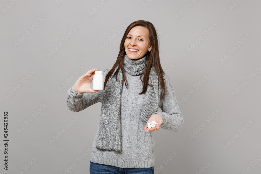 Smiling young woman in gray sweater scarf holding medication tablets, aspirin pills in bottle isolated on grey background in studio. Healthy lifestyle, ill sick disease treatment, cold season concept. Smiling young woman in gray sweater scarf holding medication tablets, aspirin pills in bottle isolated on grey background in studio. Healthy lifestyle, ill sick disease treatment, cold season concept.