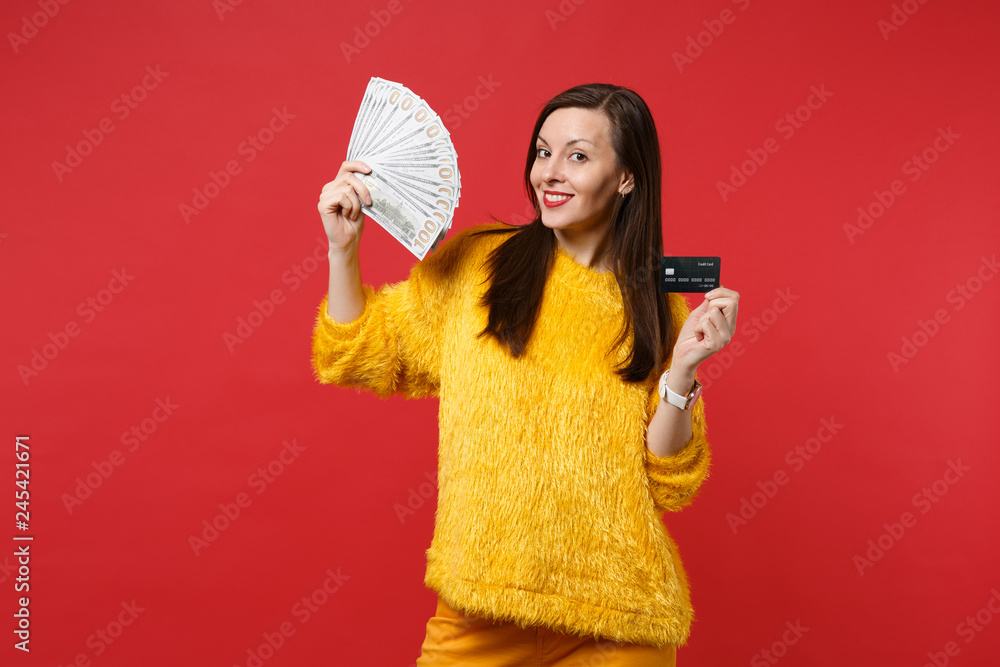Smiling young woman in yellow fur sweater hold credit bank card, fan of money in dollar banknotes, cash money isolated on red background. People sincere emotions lifestyle concept. Mock up copy space. Smiling young woman in yellow fur sweater hold credit bank card, fan of money in dollar banknotes, cash money isolated on red background. People sincere emotions lifestyle concept. Mock up copy space.