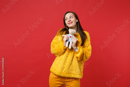 Pensive young woman in yellow fur sweater looking up, holding teddy bear plush toy isolated on bright red wall background in studio. People sincere emotions, lifestyle concept. Mock up copy space.