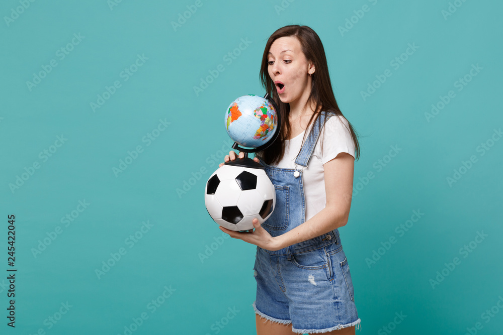 Shocked young woman football fan holding soccer ball, looking on Earth world globe isolated on blue turquoise background. People emotions, sport family leisure lifestyle concept. Mock up copy space. Shocked young woman football fan holding soccer ball, looking on Earth world globe isolated on blue turquoise background. People emotions, sport family leisure lifestyle concept. Mock up copy space.