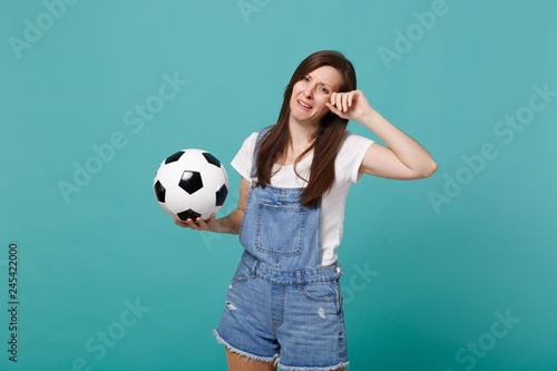 Crying distempered young woman football fan support favorite team with soccer ball wiping tears isolated on blue turquoise wall background. People emotions, sport family leisure lifestyle concept. © ViDi Studio