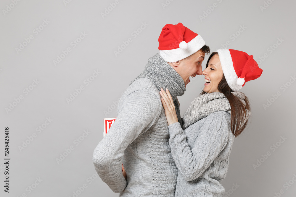 Merry fun couple girl guy in red Santa Christmas hat gray sweaters scarves isolated on grey wall background, studio portrait. Happy New Year 2019 celebration holiday party concept. Mock up copy space. Merry fun couple girl guy in red Santa Christmas hat gray sweaters scarves isolated on grey wall background, studio portrait. Happy New Year 2019 celebration holiday party concept. Mock up copy space.