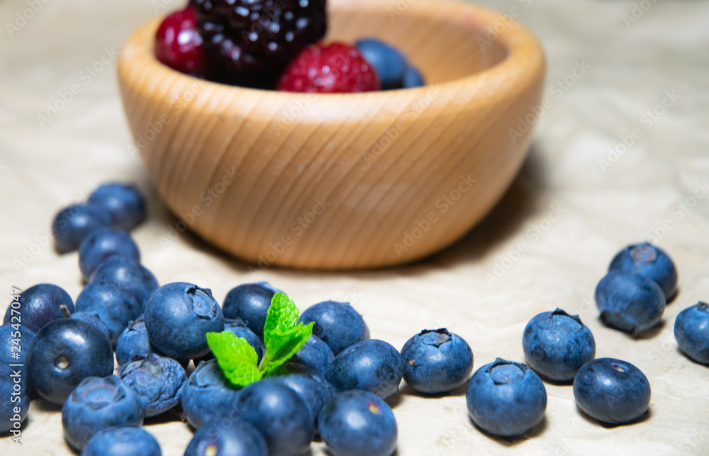 An isolated wooden bowl full of blueberries and wildberries with plain soft background decorated by fresh mint and with a few blueberries around on the paper An isolated wooden bowl full of blueberries and wildberries with plain soft background decorated by fresh mint and with a few blueberries around on the paper