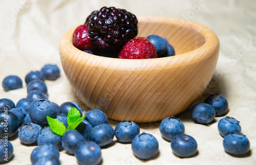 An isolated wooden bowl full of blueberries and wildberries with plain soft background decorated by fresh mint and with a few blueberries around on the paper