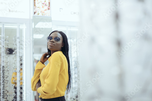 Young pretty african woman in yellow sweater choose sunglasses in Optician's shop