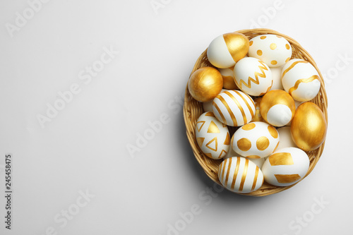 Wicker basket of traditional Easter eggs decorated with golden paint on white background, top view. Space for text