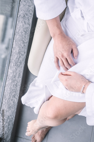 Woman sitting on man's laps in white hotel robes touching each other hands. Overhead, man's and woman's legs from above. Intimacy, Valentine's day concept