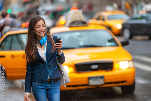 Young woman walking in New York city using phone app for taxi ride hailing with headphones commuting from work. Asian girl happy texting on smartphone. Urban walk commuter NYC.