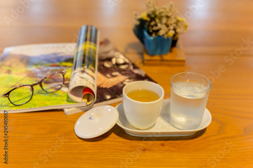Cup of tea and glasses of water with glasses with magazine on wooden table.