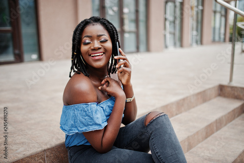 Attractive african american woman with dreads in jeans wear sitting on stairs with mobile phone.