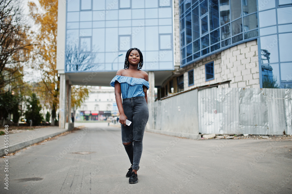 Attractive african american woman with dreads in jeans wear posed against modern multistory building.