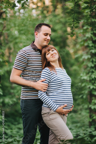 Stylish guy and his beloved pregnant girl hugging, standing in the park, against the backdrop of greenery and trees. Evening walk at sunset. First pregnancy. Love of married people.