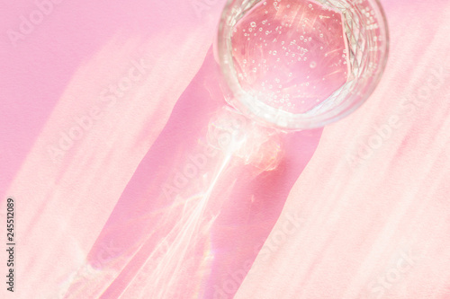 Top view of sparkling water in transparent glass and sun glare on pink background.