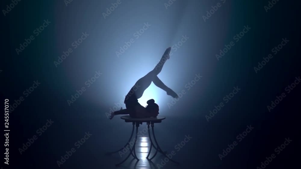 Acrobat on the table shows the tricks of standing on her hands . Smoke background. Slow motion. Silhouette Acrobat on the table shows the tricks of standing on her hands . Smoke background. Slow motion. Silhouette