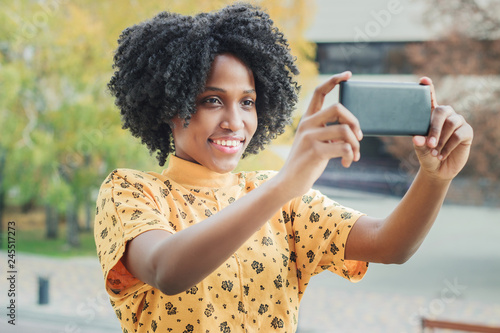 Young smiling dark-skinned woman dressed in casual clothes make a photo by her phone