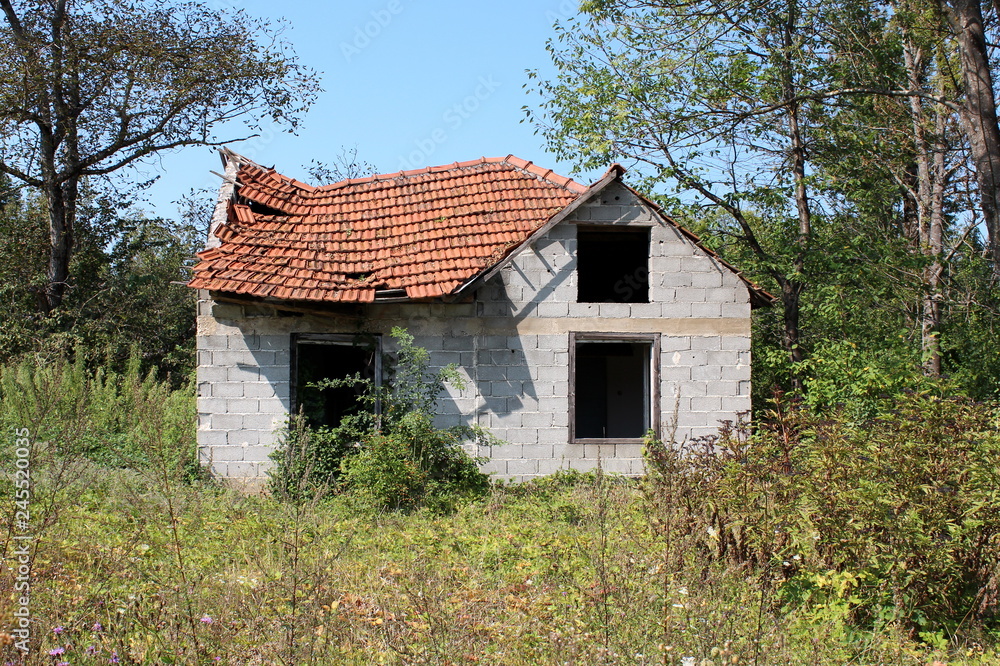 Destroyed unfinished small family house with missing windows and collapsed roof completely surrounded with overgrown vegetation and forest with clear blue sky in background Destroyed unfinished small family house with missing windows and collapsed roof completely surrounded with overgrown vegetation and forest with clear blue sky in background