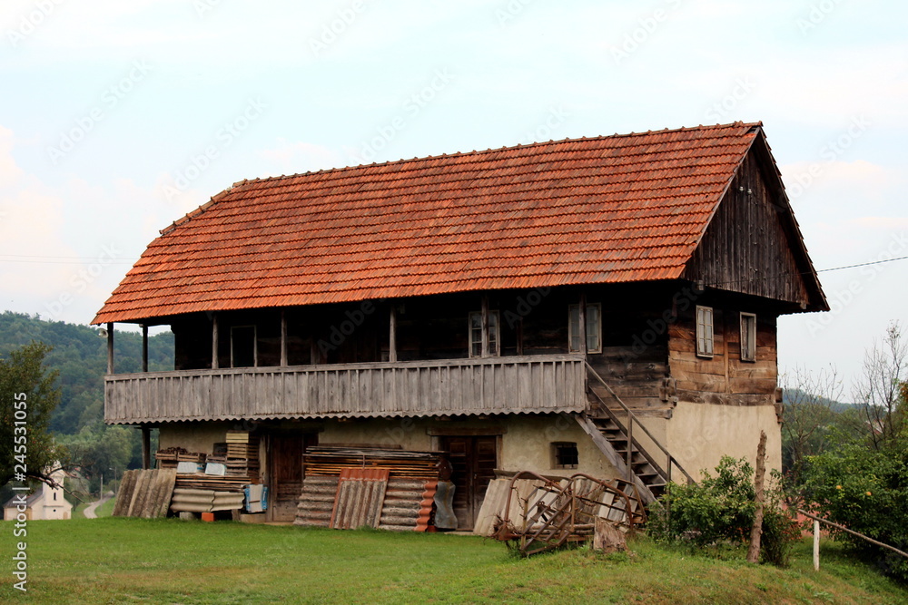 Large old wooden house with dilapidated cracked boards and windows with front porch above used tools and wooden boards covered with roof tiles surrounded with grass and forest in background