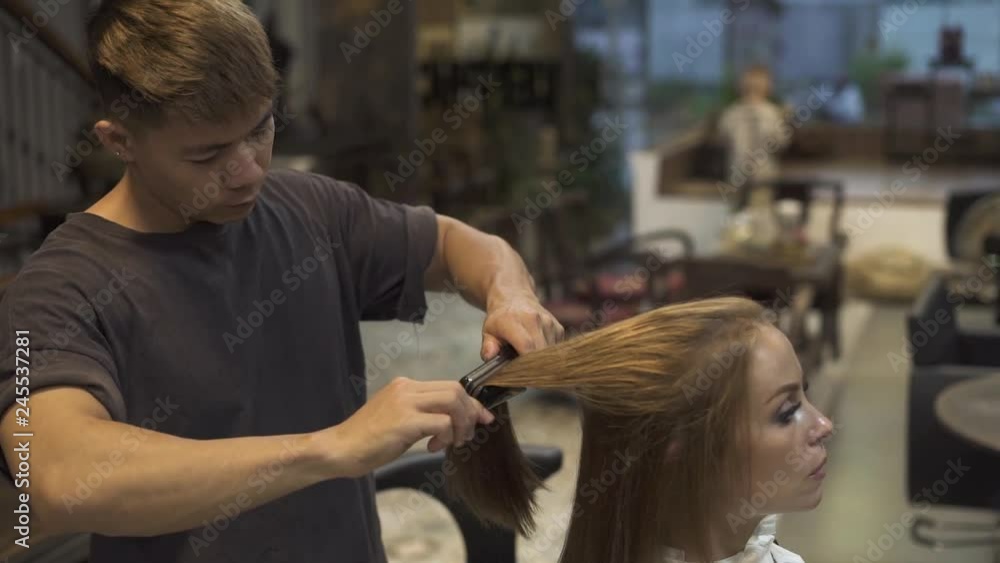 Barber using comb and hair iron for straightening hair while hairstyling in hairdressing salon. Close up female hairstyling in professional studio. Hairstylist creating woman hairstyle in barbershop. Barber using comb and hair iron for straightening hair while hairstyling in hairdressing salon. Close up female hairstyling in professional studio. Hairstylist creating woman hairstyle in barbershop.