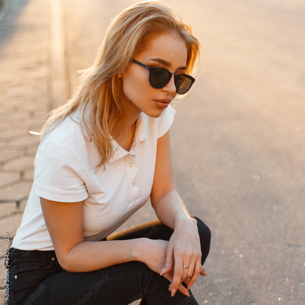 Portrait of a beautiful hipster young woman in stylish sunglasses in a  white polo t-shirt in black jeans at sunset. American girl is sitting on an  asphalt road. Stock Photo | Adobe