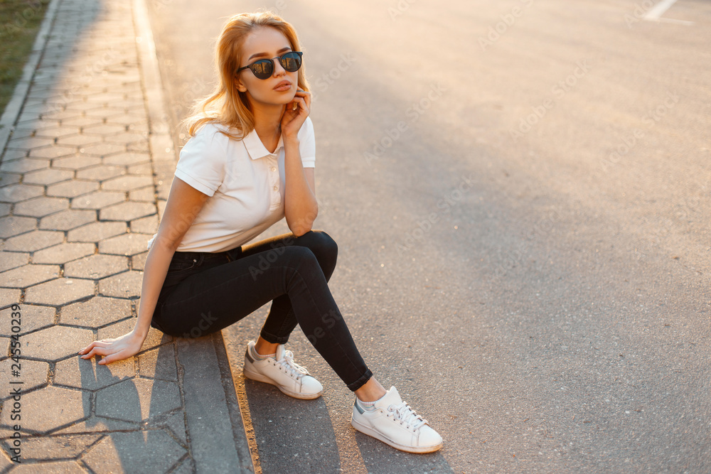 Young woman hipster in stylish sunglasses in a white polo shirt in black jeans in white fashionable sneakers sits on the asphalt against the background of a summer sunset. Pretty girl resting. - obrazy, fototapety, plakaty 