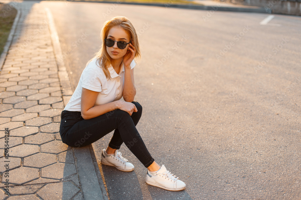 Young beautiful stylish hipster woman with sunglasses in a white polo shirt  and black jeans with sneakers sitting on the street at sunset. Fashionable  girl in summer clothes Stock Photo | Adobe