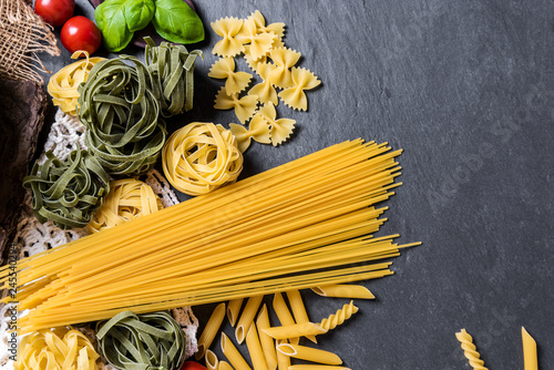 Assortment of many different raw pasta on a rustic board, beautifully arranged with basil and tomatoes