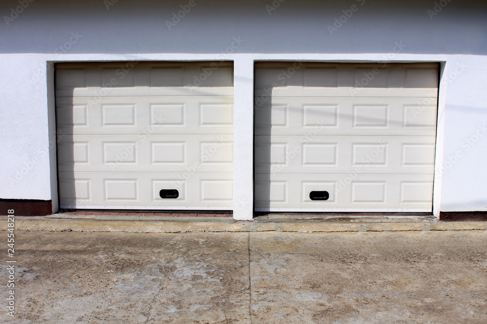 Two modern white rollup garage doors with cracked concrete driveway mounted on white wall on warm sunny day Two modern white rollup garage doors with cracked concrete driveway mounted on white wall on warm sunny day