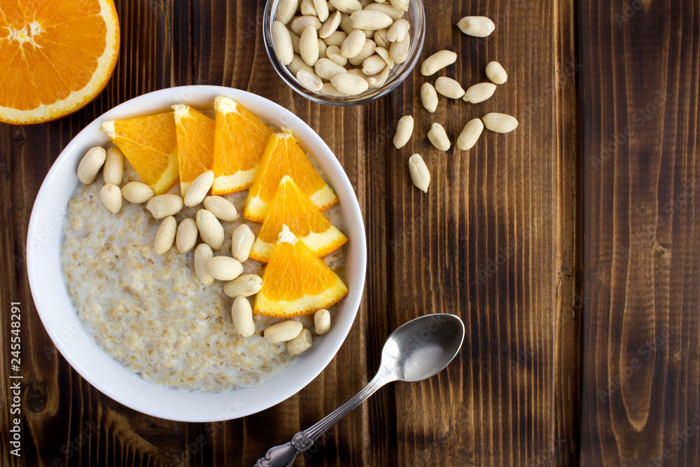 Breakfast: oatmeal with orange and peanuts in the white bowl on the brown wooden background.Top view.Copy space. Breakfast: oatmeal with orange and peanuts in the white bowl on the brown wooden background.Top view.Copy space.