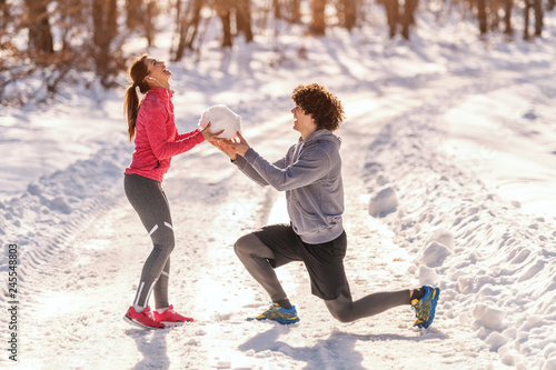 Runners on the snow. Man kneeling and giving to woman big snowball while woman laughing. Wintertime, healthy lifestyle concept.