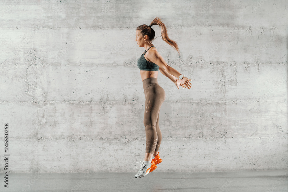 Φωτογραφία Side view of muscular focused brunette with ponytail and in sportswear jumping in front of brick wall in gym