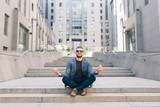 Full-length photo of  man sitting on concrete stairs on office building background. He wears T-shirt, jacket,  jeans, sunglasses. He has a fun showing meditation.