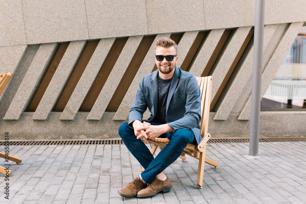 Handsome guy is sitting on chair outdoor on cafe background. He wears gray jacket, jeans, brown shoes, sunglasses. He is smiling to the camera.