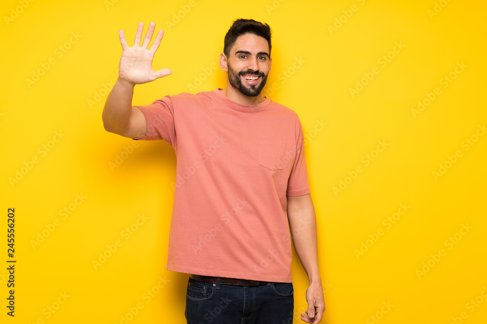 Handsome man over yellow wall saluting with hand with happy expression