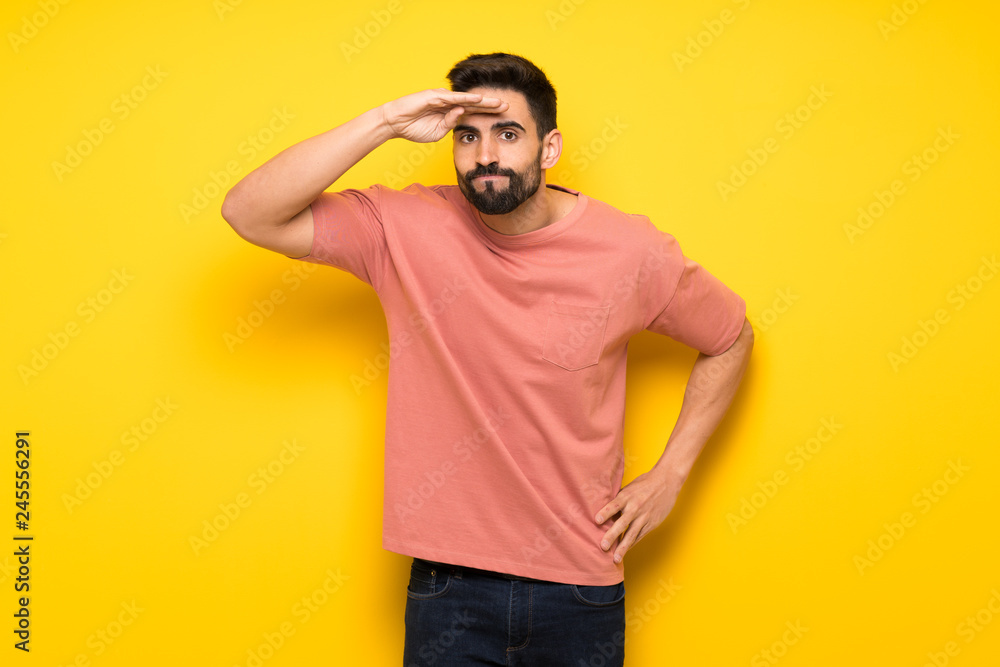 Handsome man over yellow wall looking far away with hand to look something Handsome man over yellow wall looking far away with hand to look something